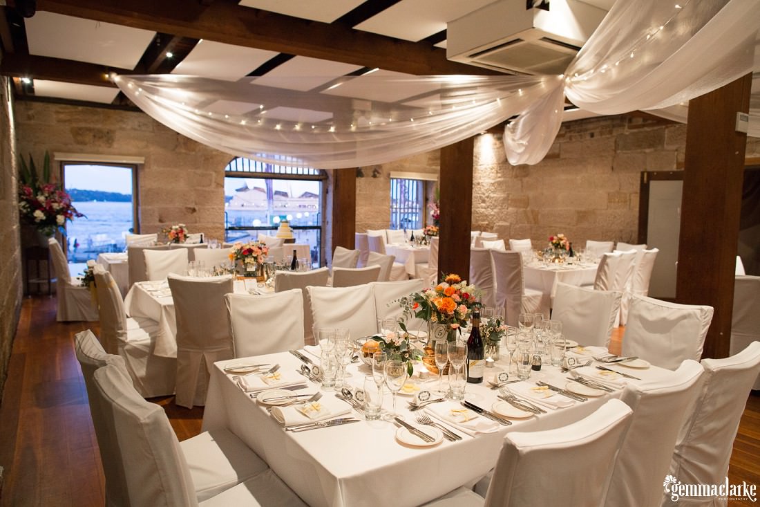 An intimate wedding reception setup with white tables and chair covers, white ceiling draping and floral centrepieces in a room with sandstone walls - The Rocks Wedding