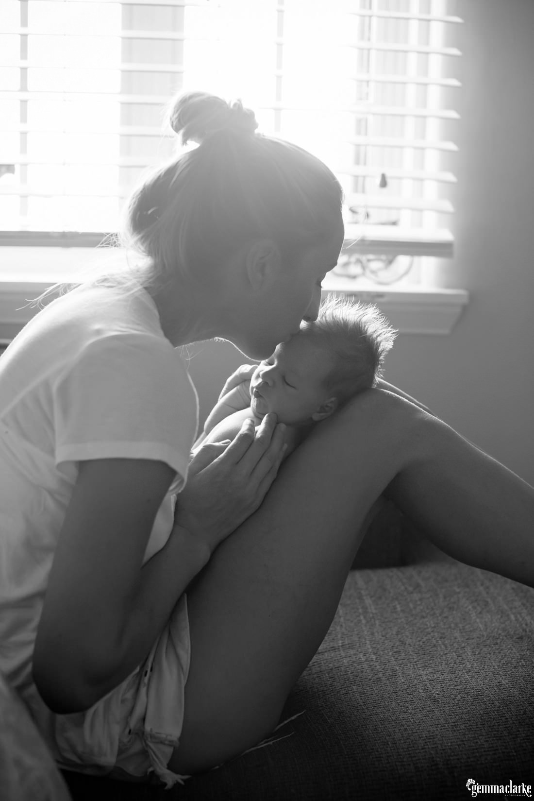 A mother with her newborn son in her lap, kissing him on the forehead - Newborn Lifestyle Portraits