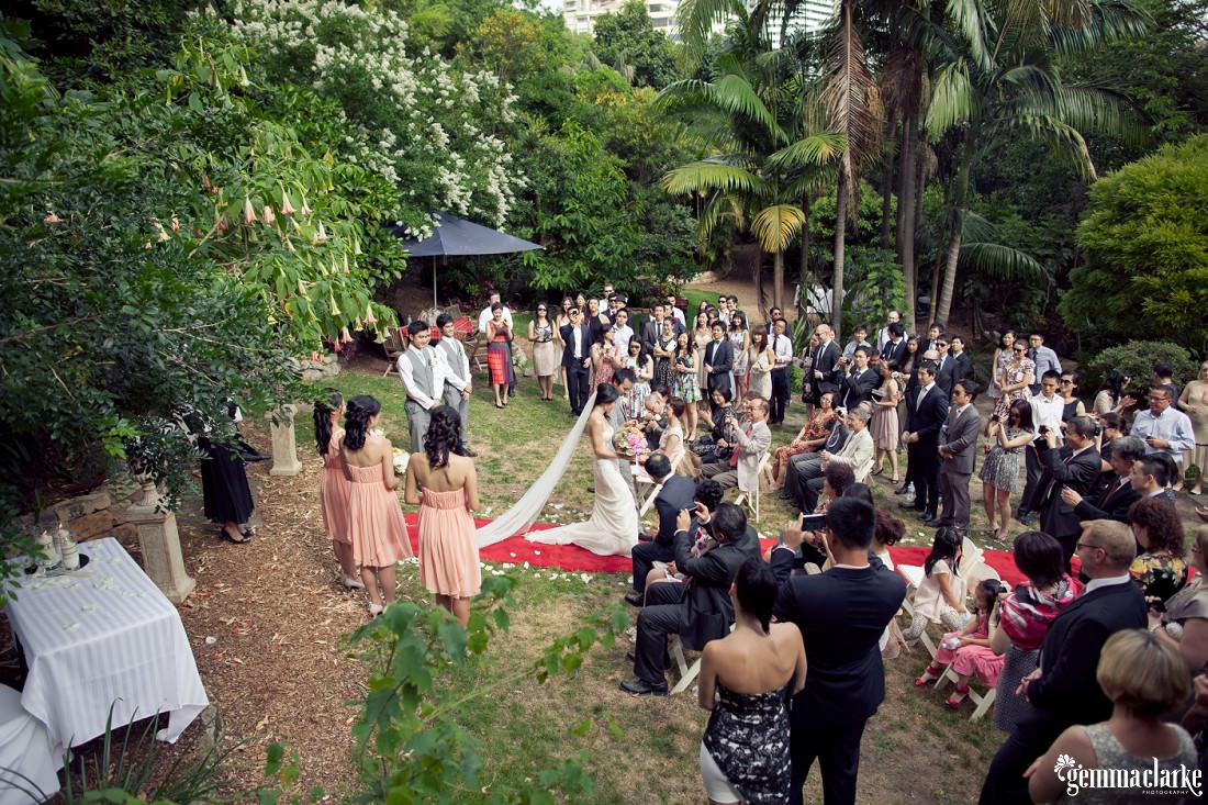 A higher view of the ceremony in a garden with lots of guests sitting and standing and the bride and groom walking down the red carpet - Secret Garden Wedding