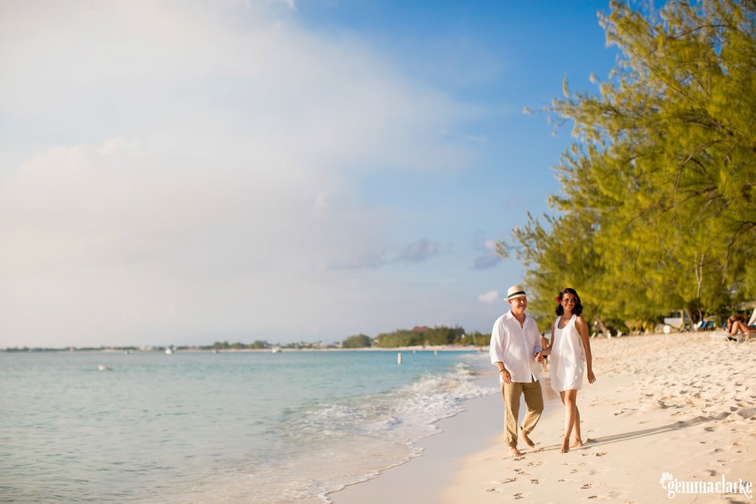Bride and groom in their casual wedding attire walking along the white sands of the cayman islands with the blue sky and water in the background at this caribbean elopement