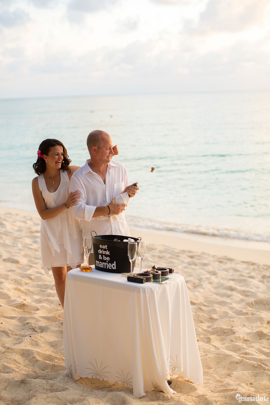 Action shot of Lee popping the champagne bottle while standing in front of a white table with champagne glasses on it at this caribbean elopement