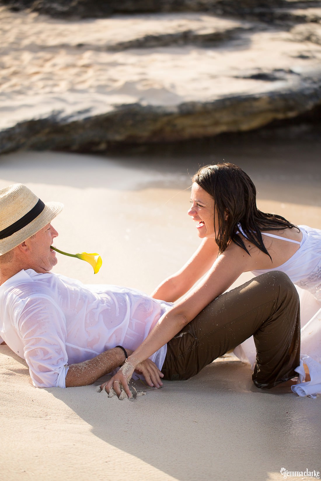 Lee lying down on the beach with a flower in his mouth enticing Marie to give him a kiss as the waves make their way up to them in this trash the dress session