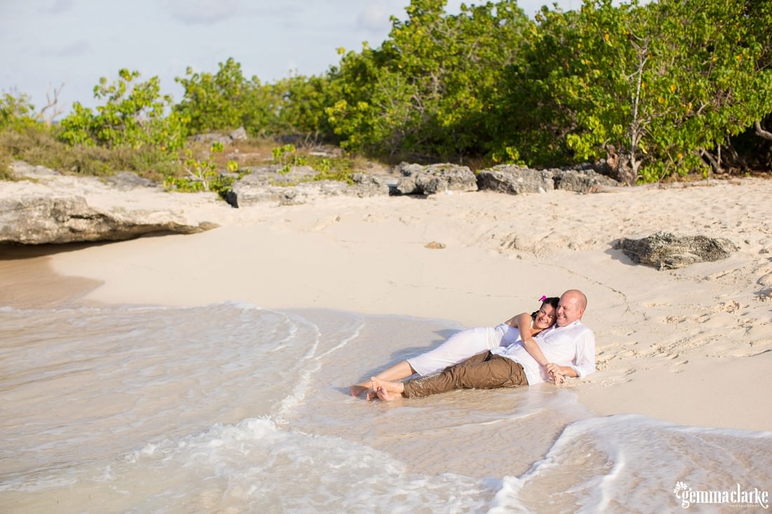 A bride and groom lay in the shallow water on the beach - Trash the Dress