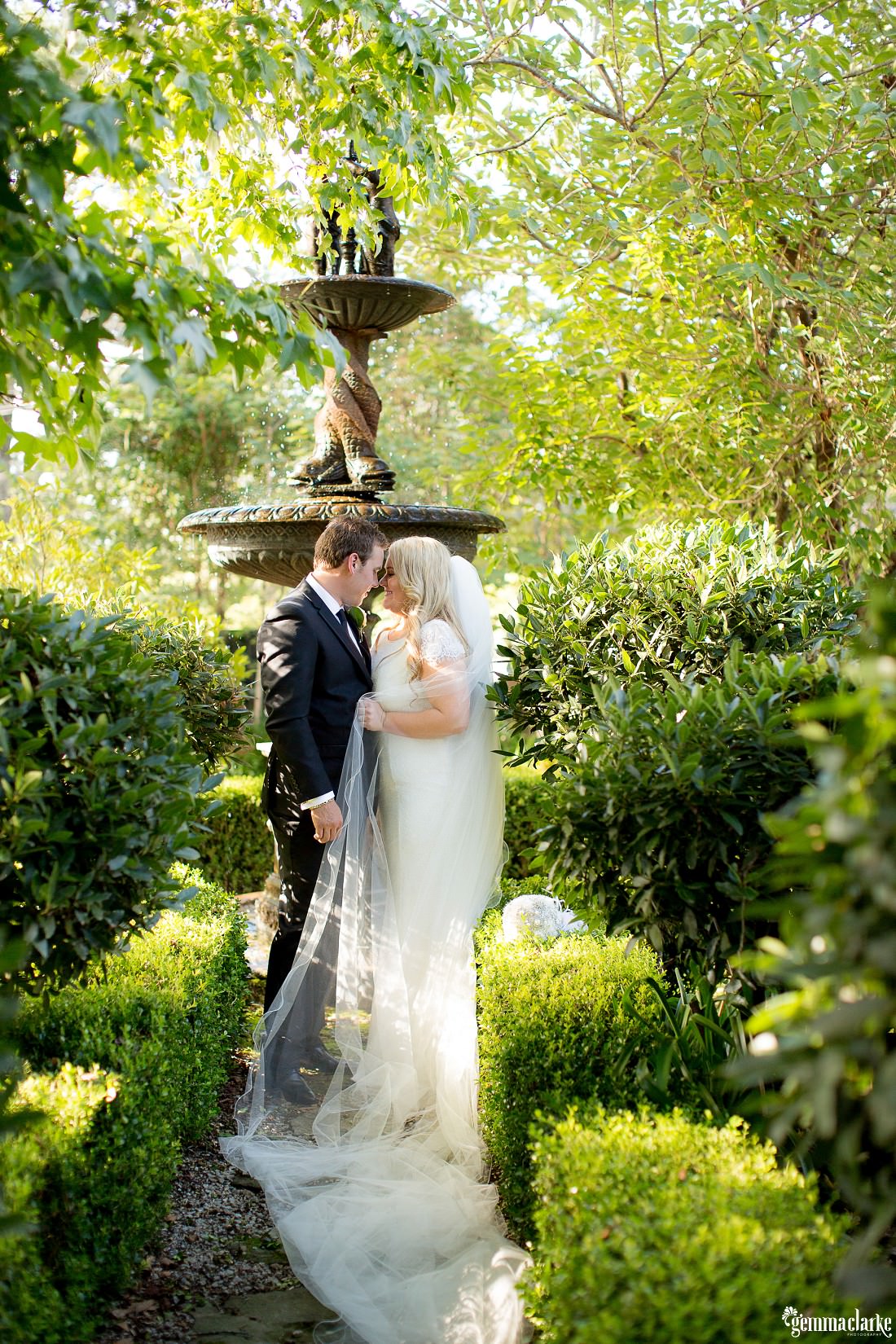 A bride and groom share an intimate moment in front of a fountain in a lush green garden - Echoes Hotel Wedding