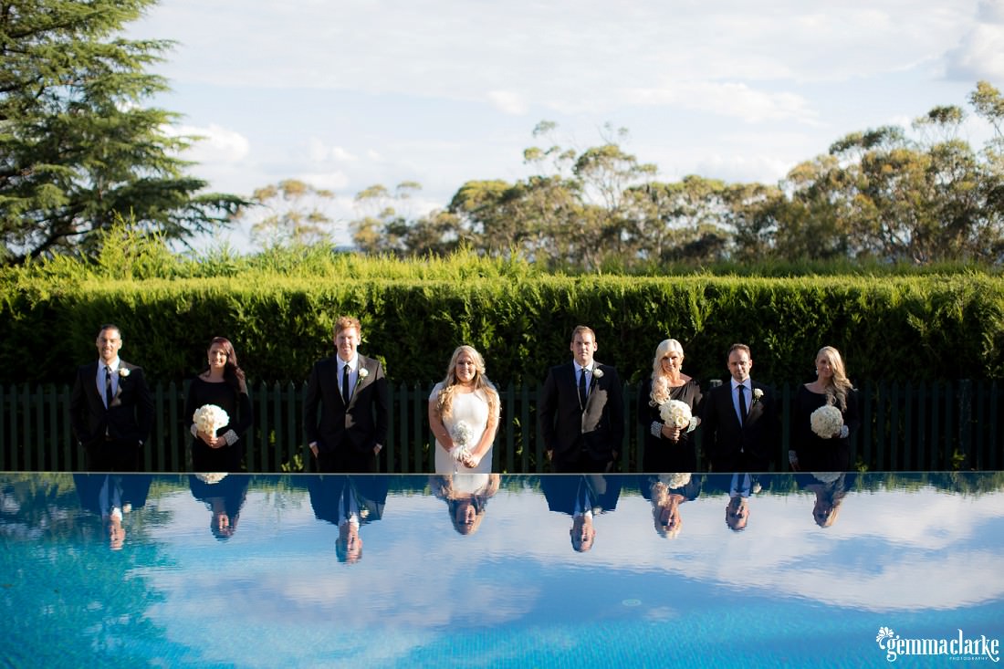 A bridal party lined up in front of a hedge with their reflections in view in a pool of water in front of them - Echoes Hotel Wedding