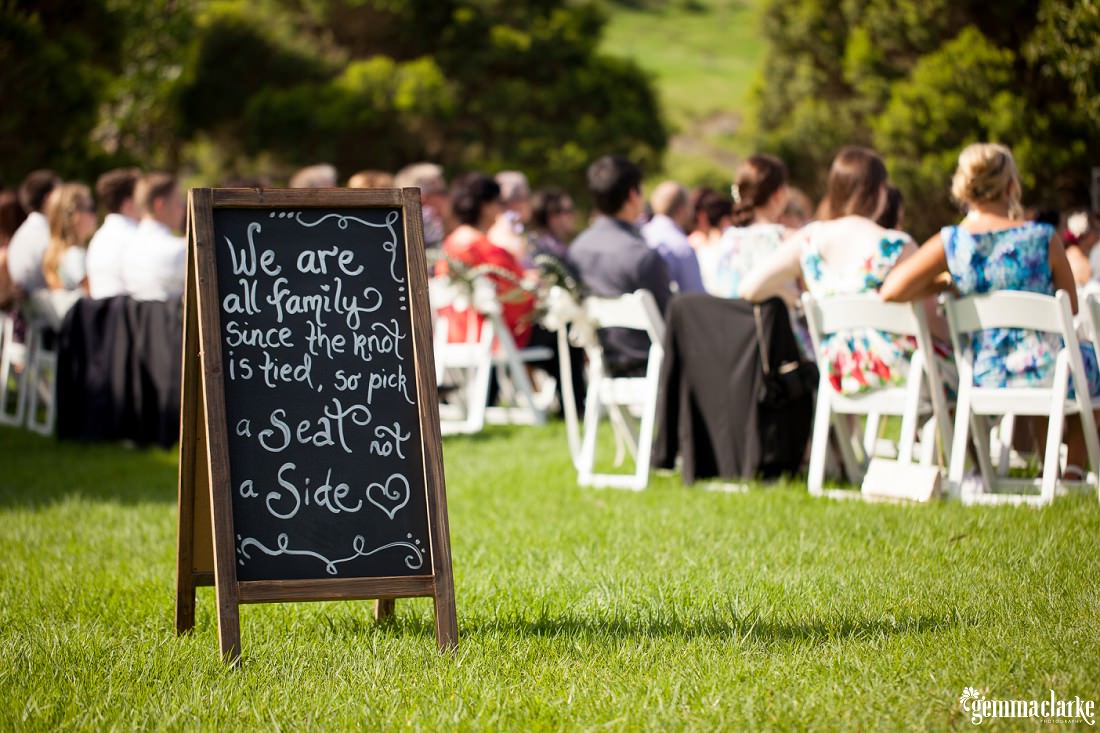 Chalkboard sign in front of guests seated at the grassy ceremony on the Hillside at this Bush Bank Wedding.