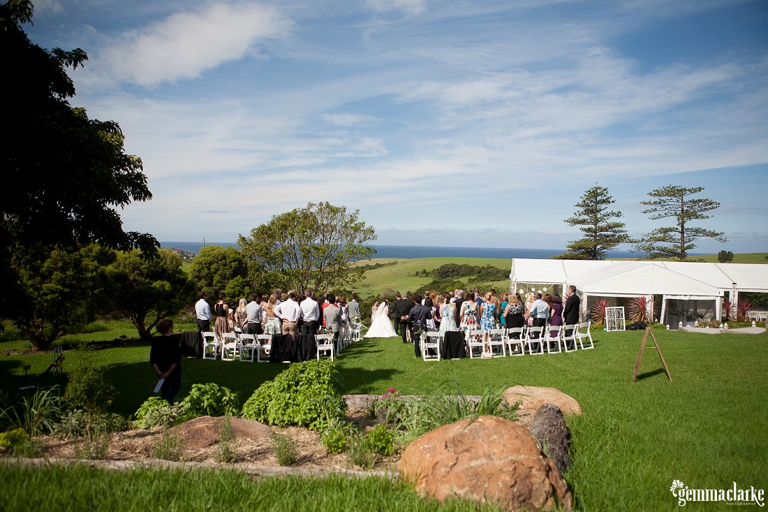 Overall view of the bush bank wedding ceremony from the back with bride and groom at the end of the aisle and a view over the grassy hills and ocean