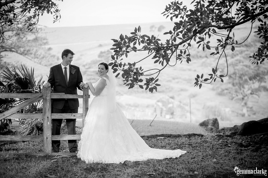 A bride and groom standing either side of a wooden fence on a hillside - Bush Bank Wedding