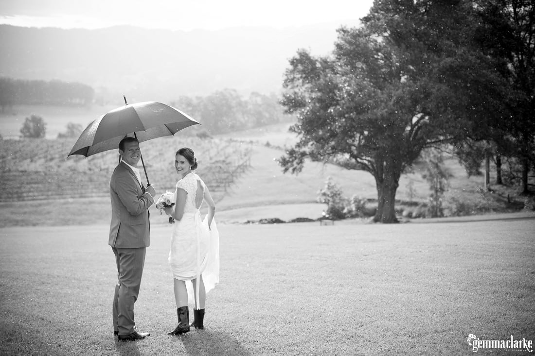 Black and White image of bride and groom walking toward the vines in the distance. The groom is holding an umbrella and the bride is wearing gumboots and holding her dress up. This Silos Estate Wedding.