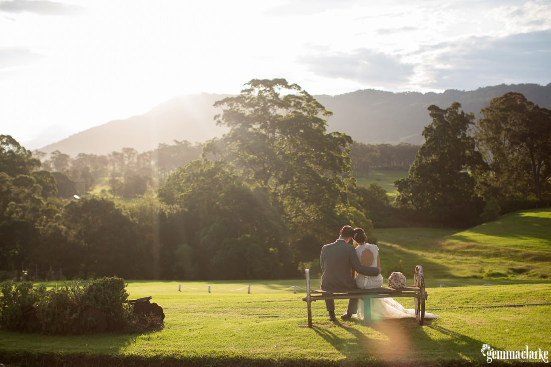 Bride and Groom sitting on an old wagon wheel type seat and looking at the sun setting over the green rolling hills at this Silos Estate Wedding