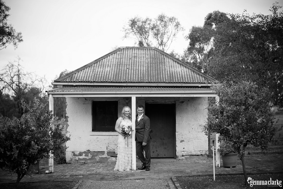 Black and white photo of a bride and groom standing in front of a small heritage cottage and leaning on a pole at this Gledswood Homestead Wedding
