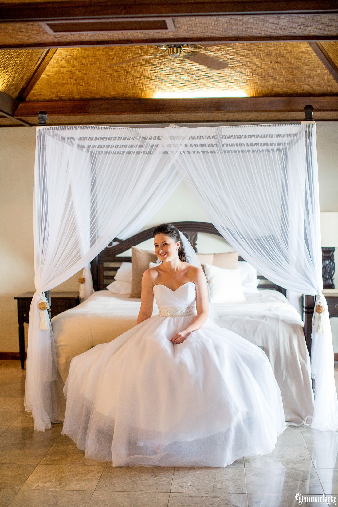 A smiling bride in her bridal gown sitting on a four poster bed with white canopy - South Pacific Wedding