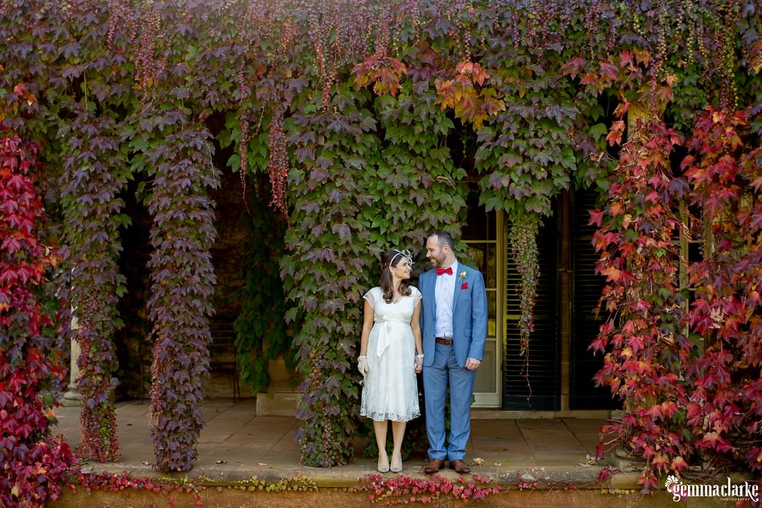 Bride and Groom holding hands in front of a vine covered building with amazing autumn colours in the leaves at this Autumn Southern Highlands Wedding