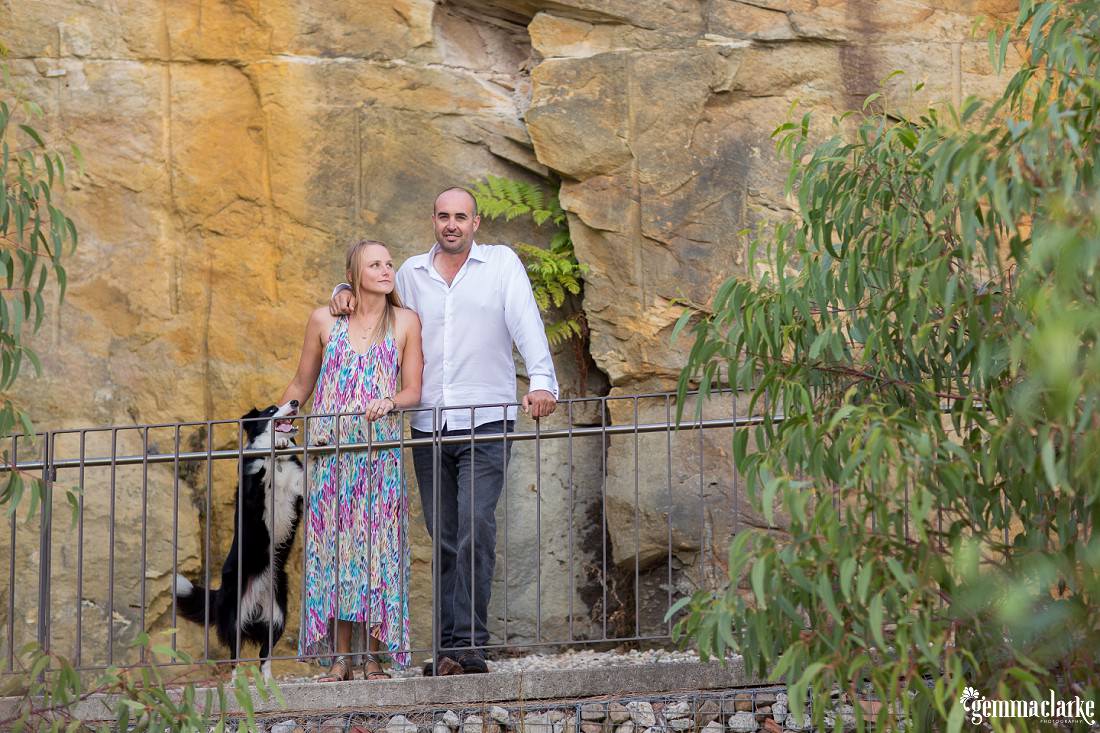 A man and woman and their dog standing together leaning against a railing in front of a large stone wall - Ballast Point Park Portraits