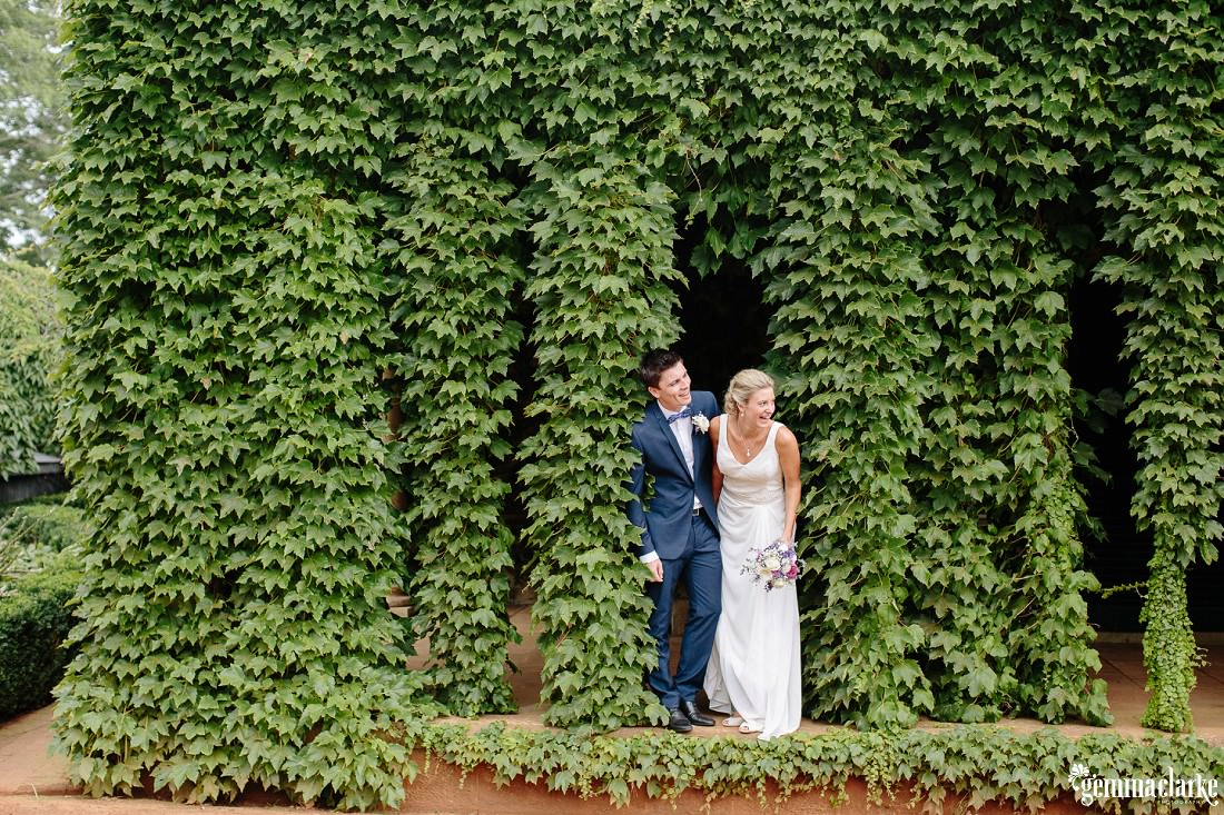 A bride and groom peeking out from between some hedges - Book Barn Wedding
