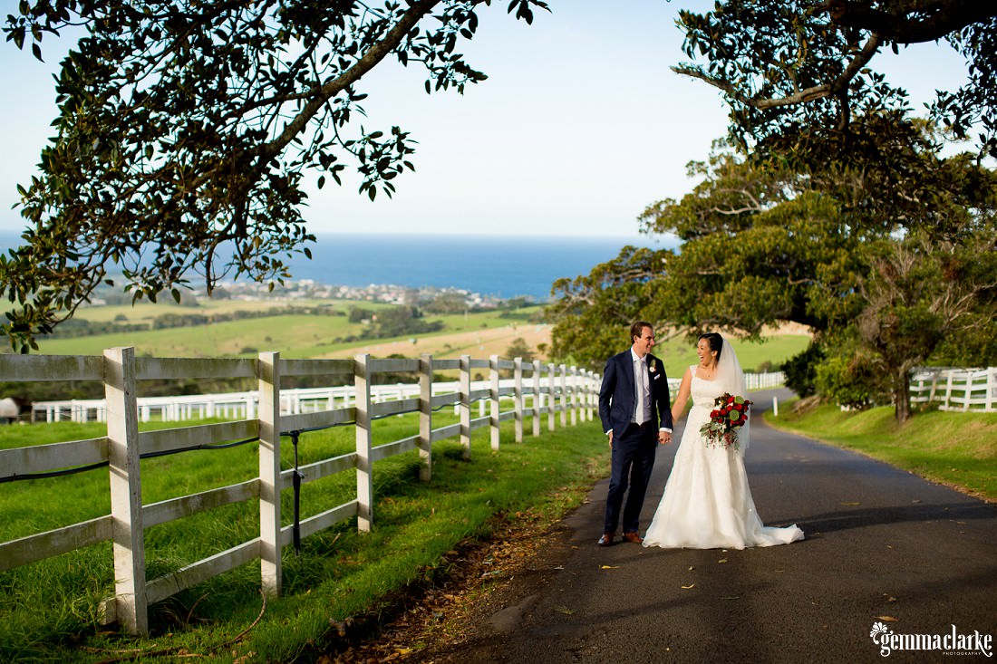 A bride and groom holding hands on a road next to a white fence with the sea in the background - Kiama Wedding