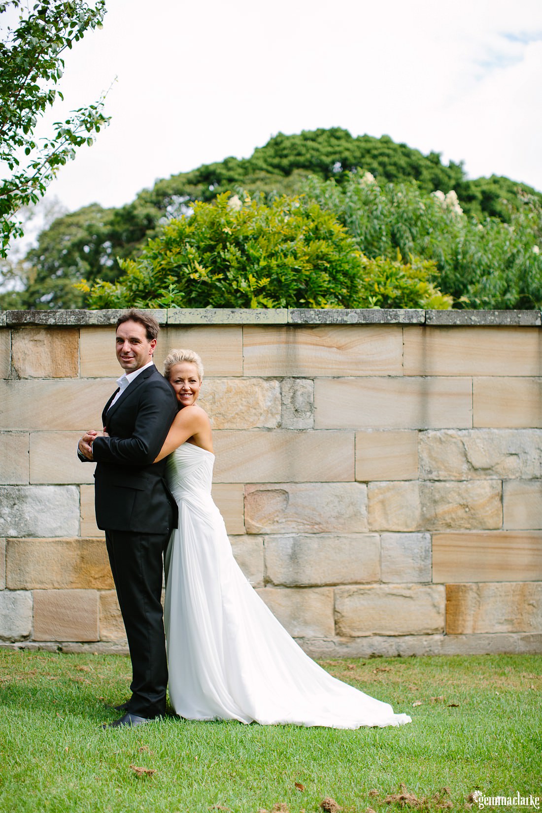 A bride stands behind her groom and holds him around the waist as they stand in front of a sandstone wall - Lion Gate Lodge Wedding