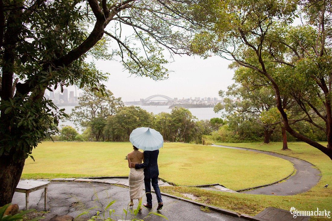A bride and groom walking together under a pale blue umbrella with Sydney Harbour in the background - Rainy Athol Hall Wedding