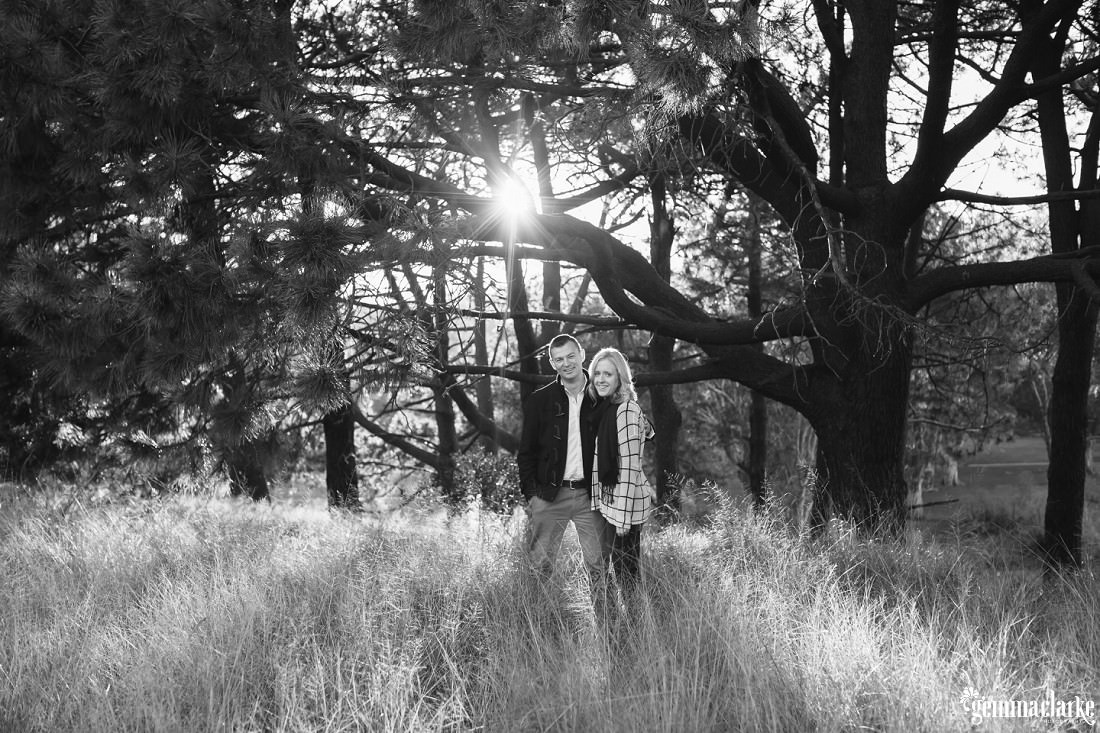 A man and woman standing in some long grass with sunlight streaming through the trees behind