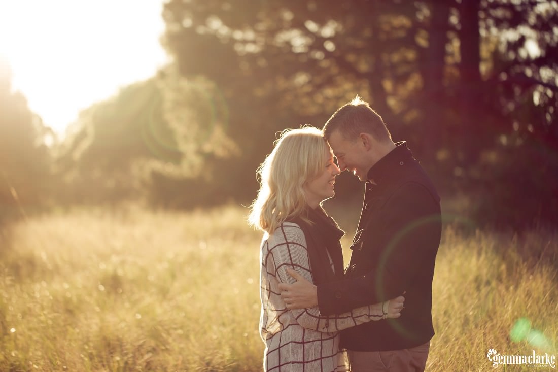 A man and woman holding each other close with their foreheads together and smiling as the sun streams in from the background and shines over the long grass