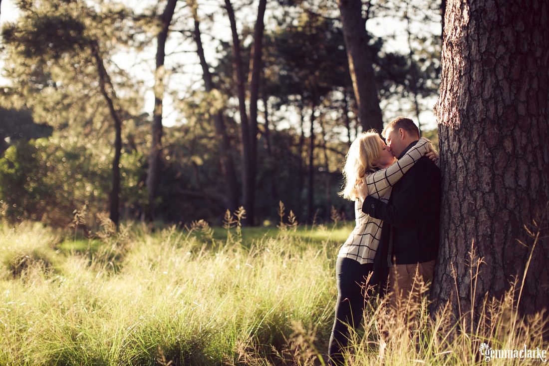 A man and woman kiss as he leans back against a tree in a field of long grass