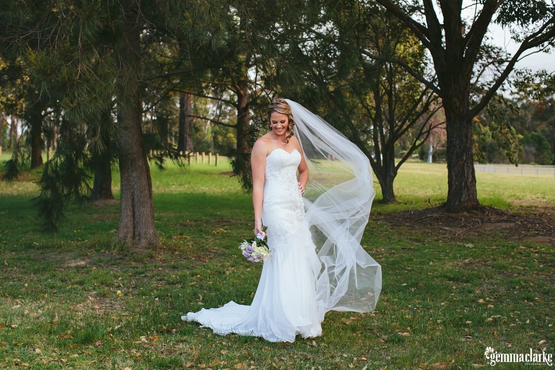 A bride poses in front of some trees, her veil blowing in the wind