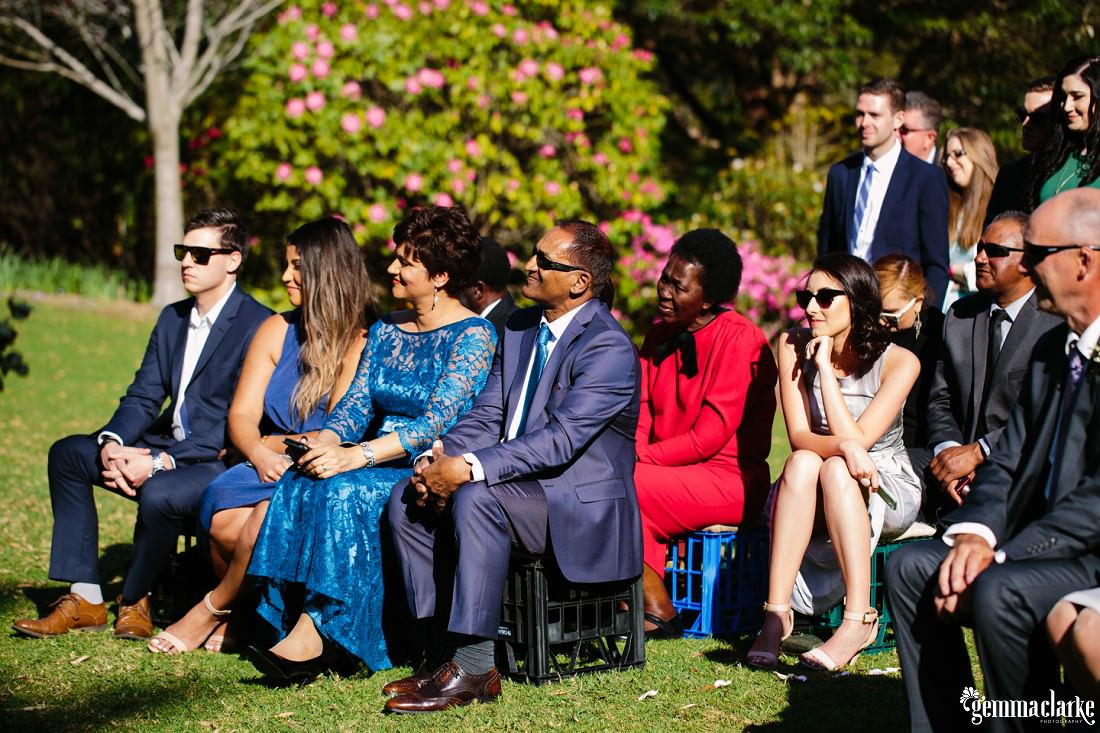 Smiling wedding guests watching a ceremony while seated on milk crates