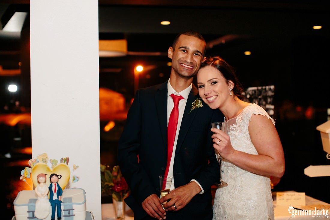 A smiling bride posing with her head on her groom's shoulder and a champagne flute in her hand, standing next to their cake
