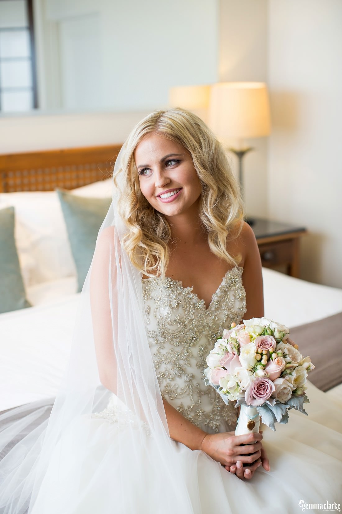 A smiling bride holding her floral bouquet and sitting on the end of a bed