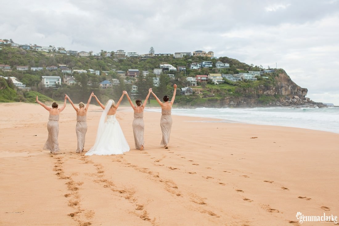 A bride and her bridesmaids standing on a beach with their hands held in the air