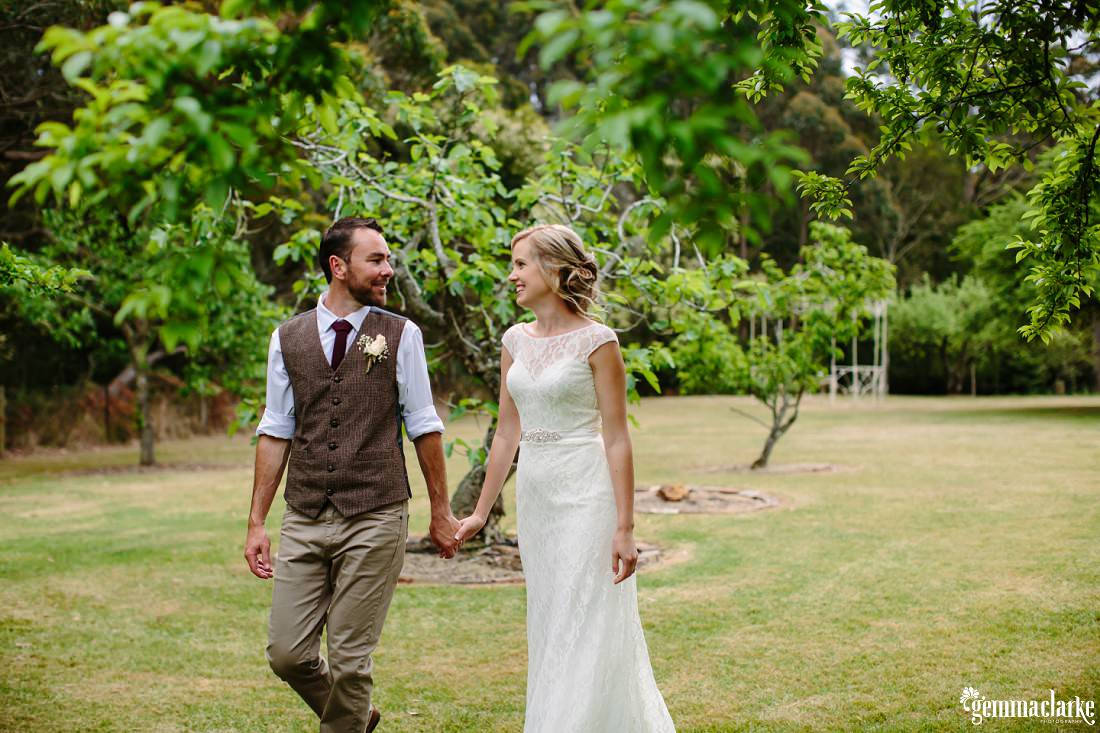 A bride and groom look at each other lovingly while walking across a lawn hand in hand