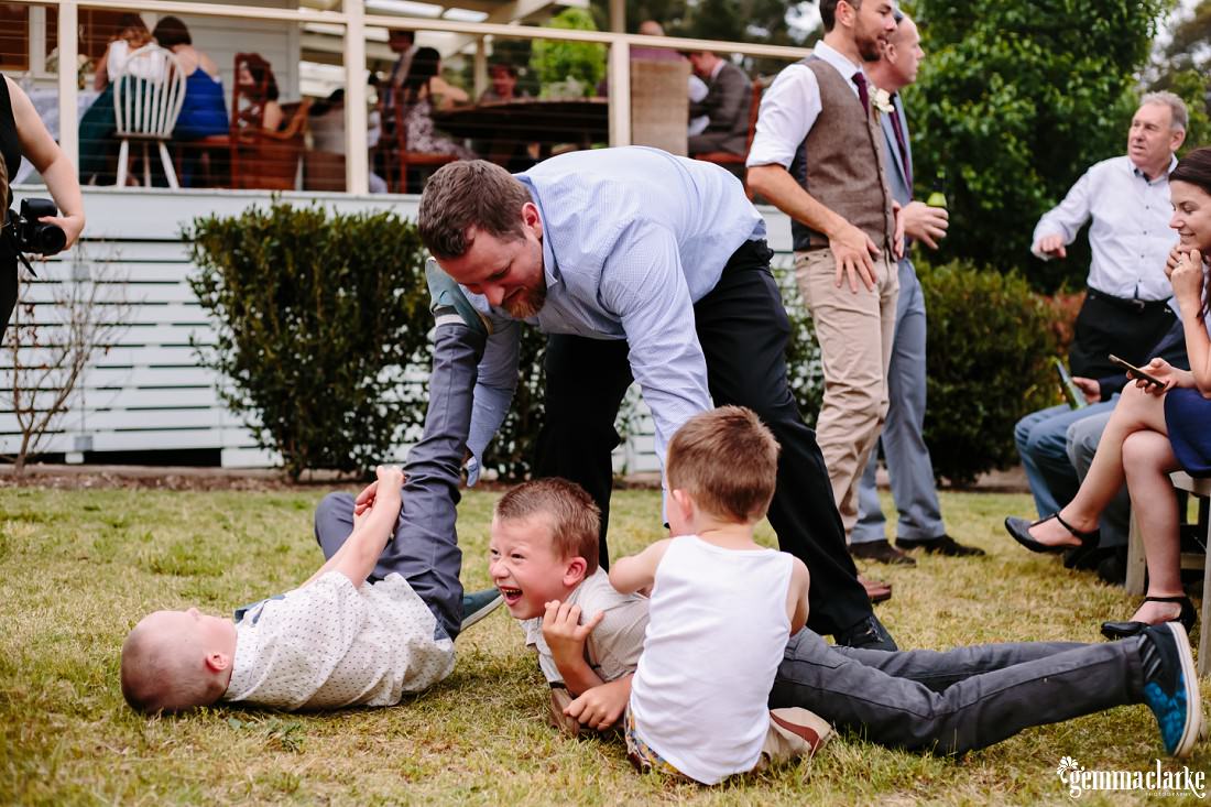 A man and three boys playing on the grass as wedding guests talk and mingle in the background