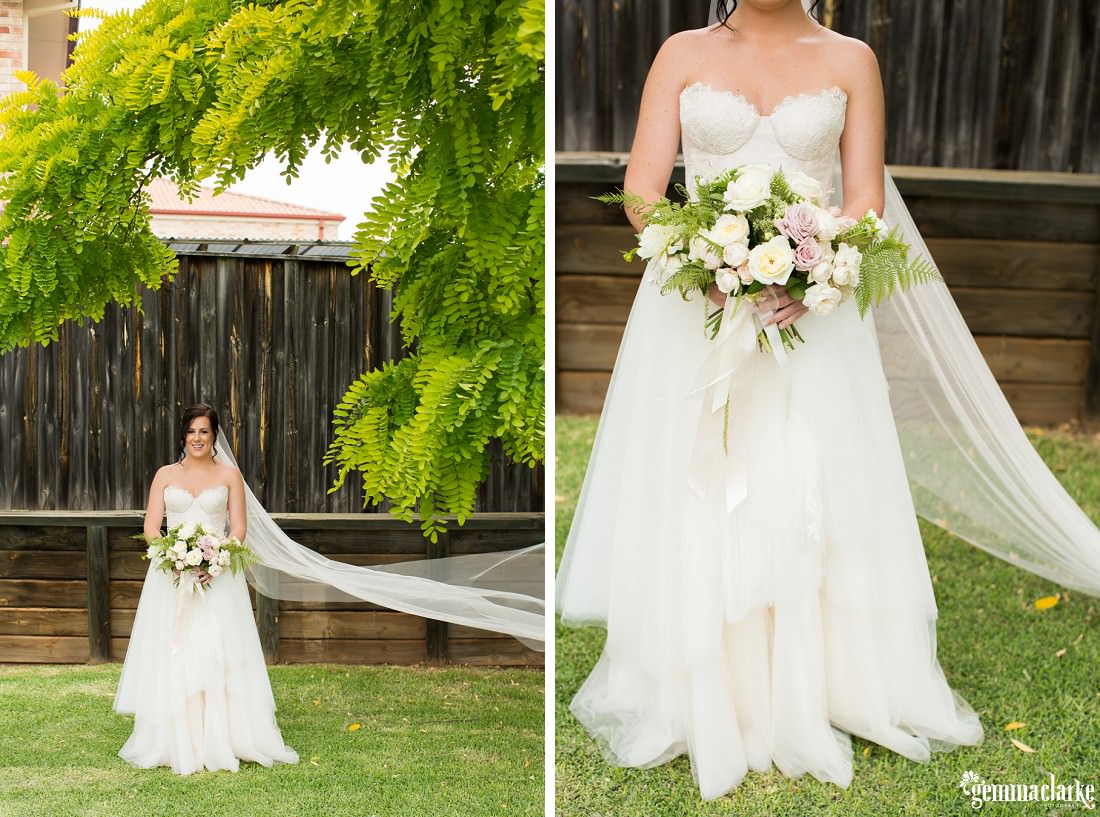 A bride holding her floral bouquet standing underneath a tree in front of a wooden fence