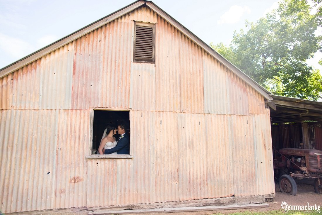 A bride and groom embracing and sharing an eskimo kiss as seen through a window in a corrugated shed