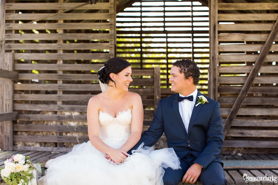 A bride and groom smile at each other and hold hands while sitting on the deck of a small wooden building
