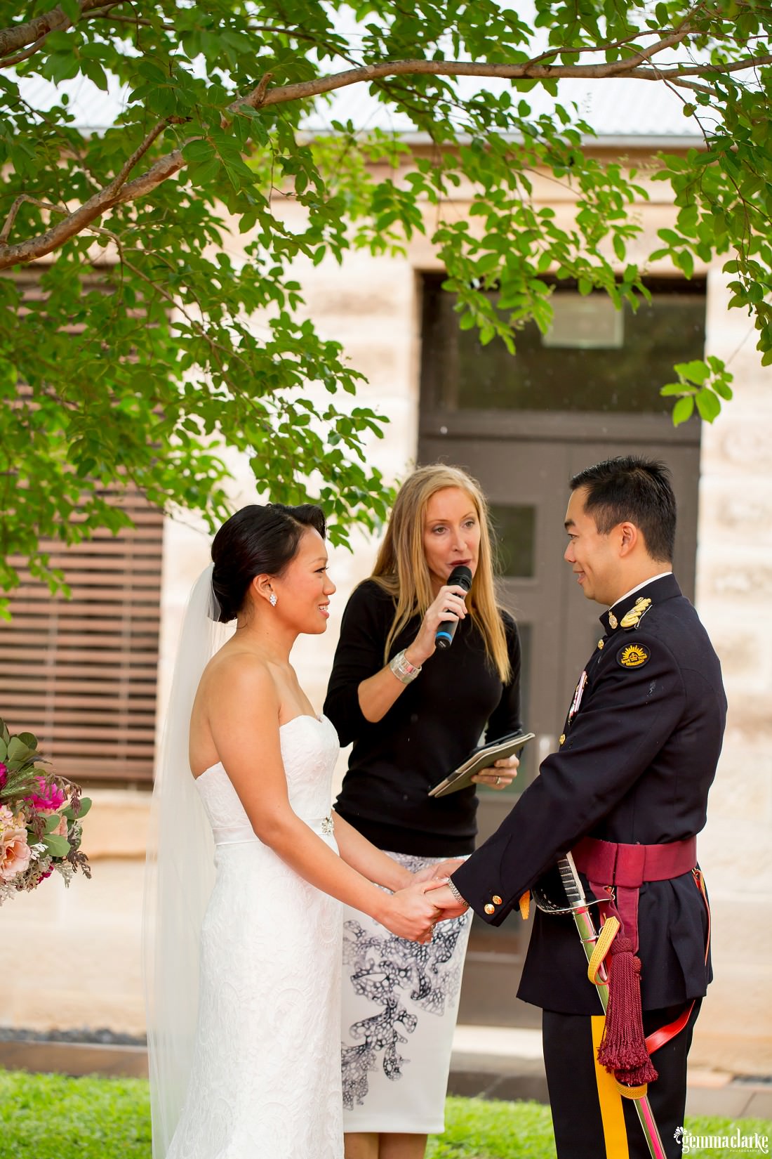 A bride and groom smile at each other and hold hands as the celebrant speaks into a microphone