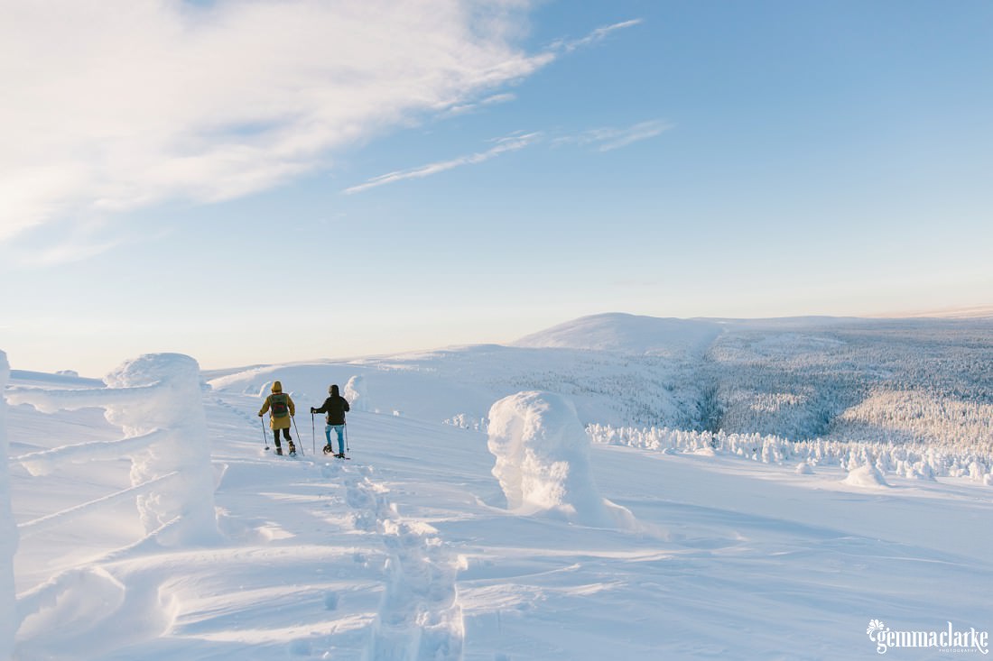 A couple snow-shoeing across a hill looking down over a snow covered forest