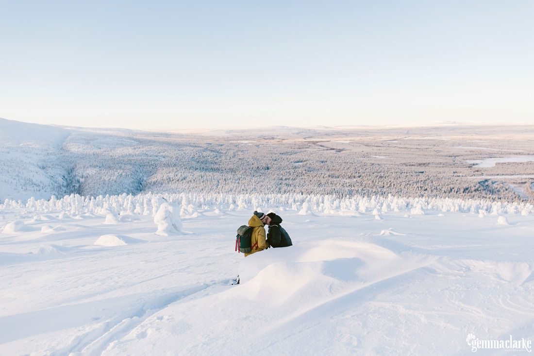 A couple kissing on a snowy hill overlooking a snow covered forest