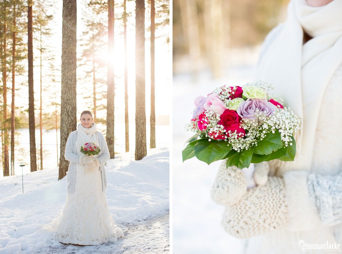 A bride in a white gown with scarf and mittens stands in the snow holding a floral bouquet