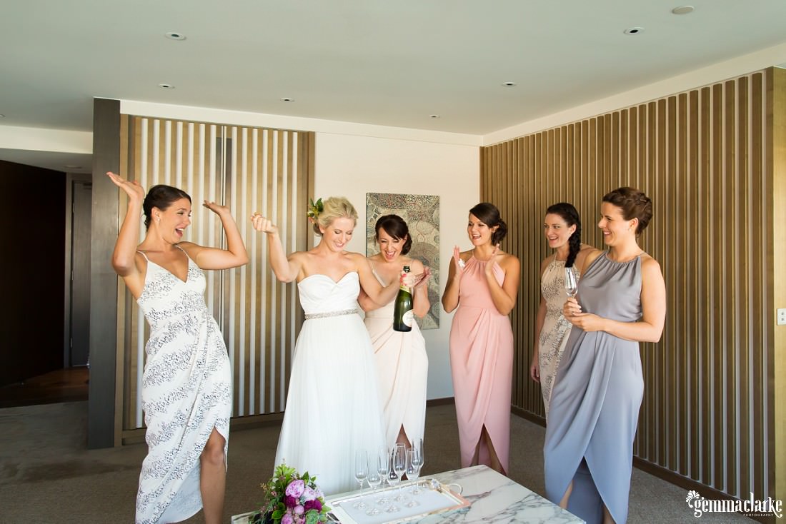 A group of bridesmaids smiling and celebrating as the bride opens a bottle of champagne