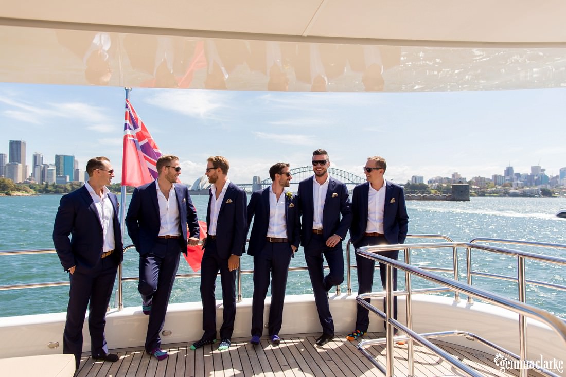 A groom and his groomsmen stand smiling at the rear of a boat with the Sydney Harbour Bridge in the background