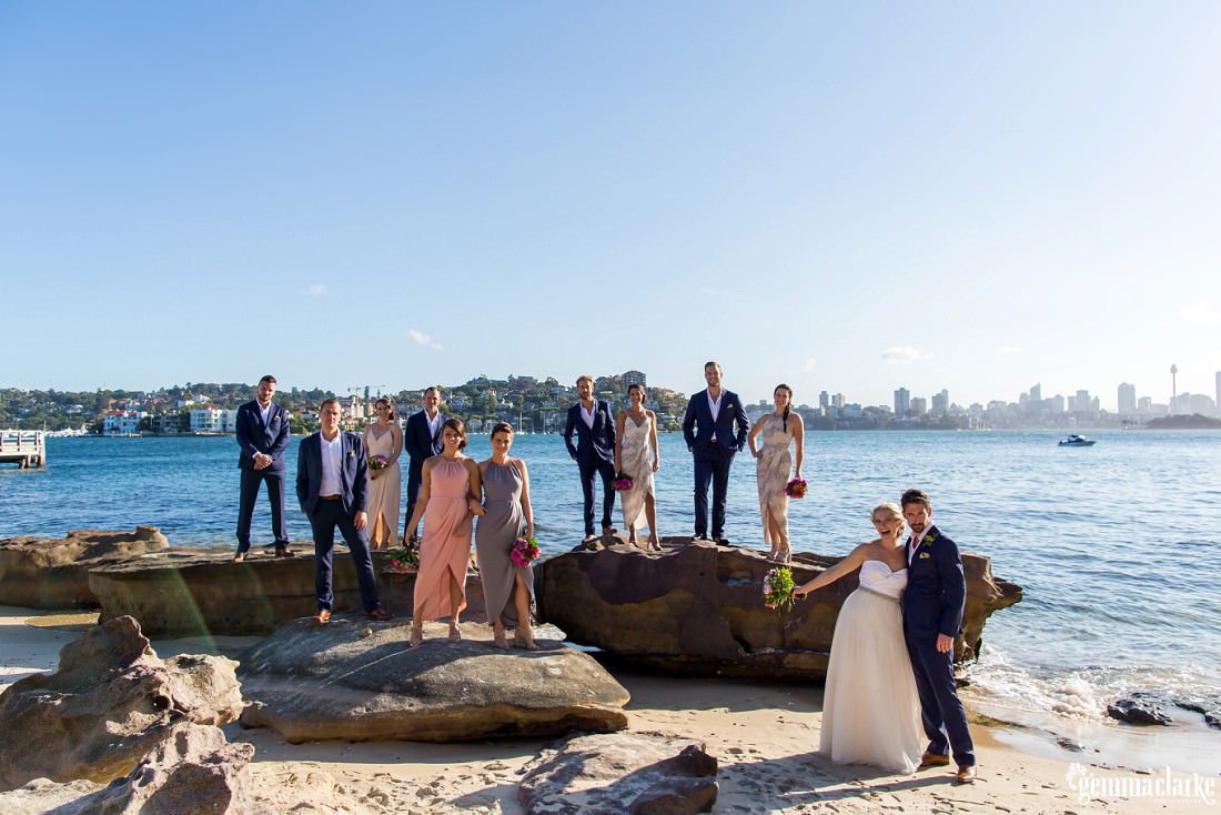 A bride and groom stand in the sand as their wedding party stands on large rocks on a beach