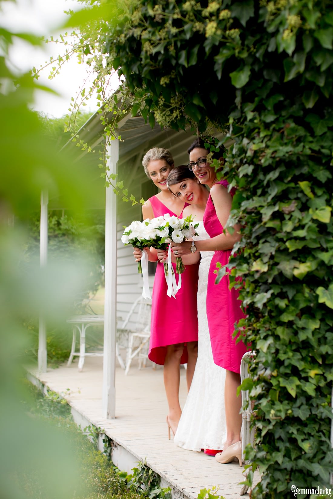 A bride and her bridesmaids standing on a verandah looking out from around a tree
