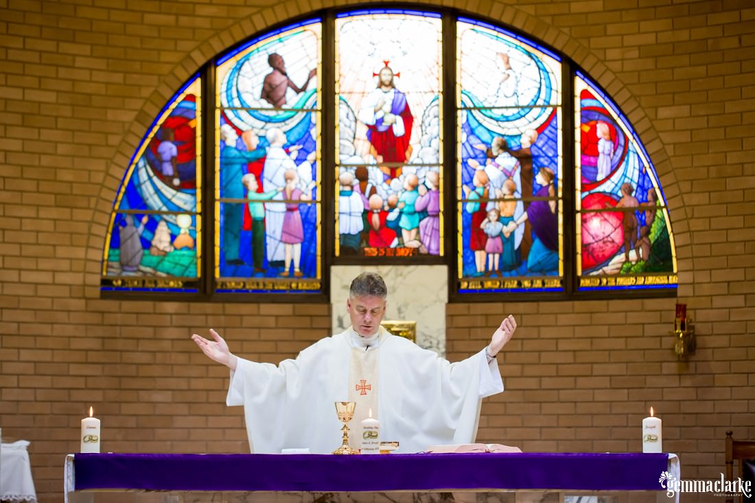 A minister with arms outstretched with stained glass in the background
