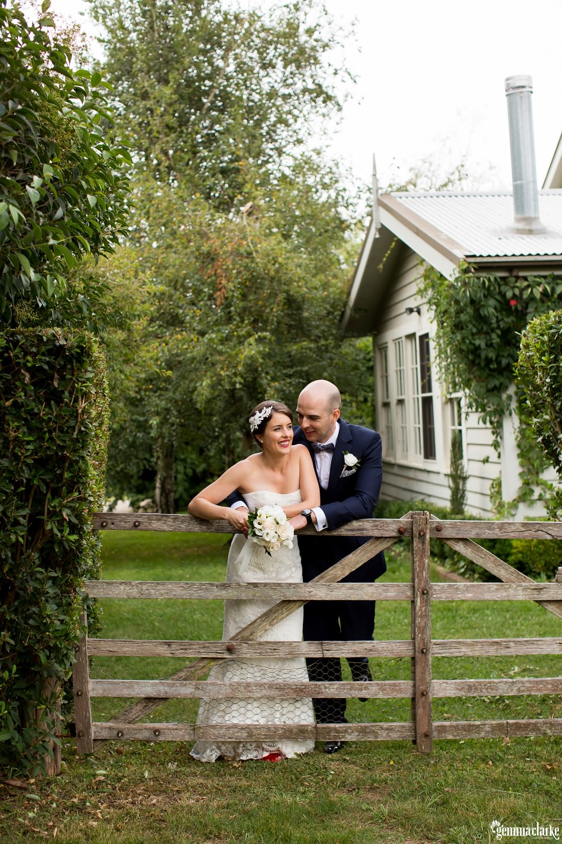 A groom embraces his smiling bride as they lean on a wooden country gate after their Summerlees wedding