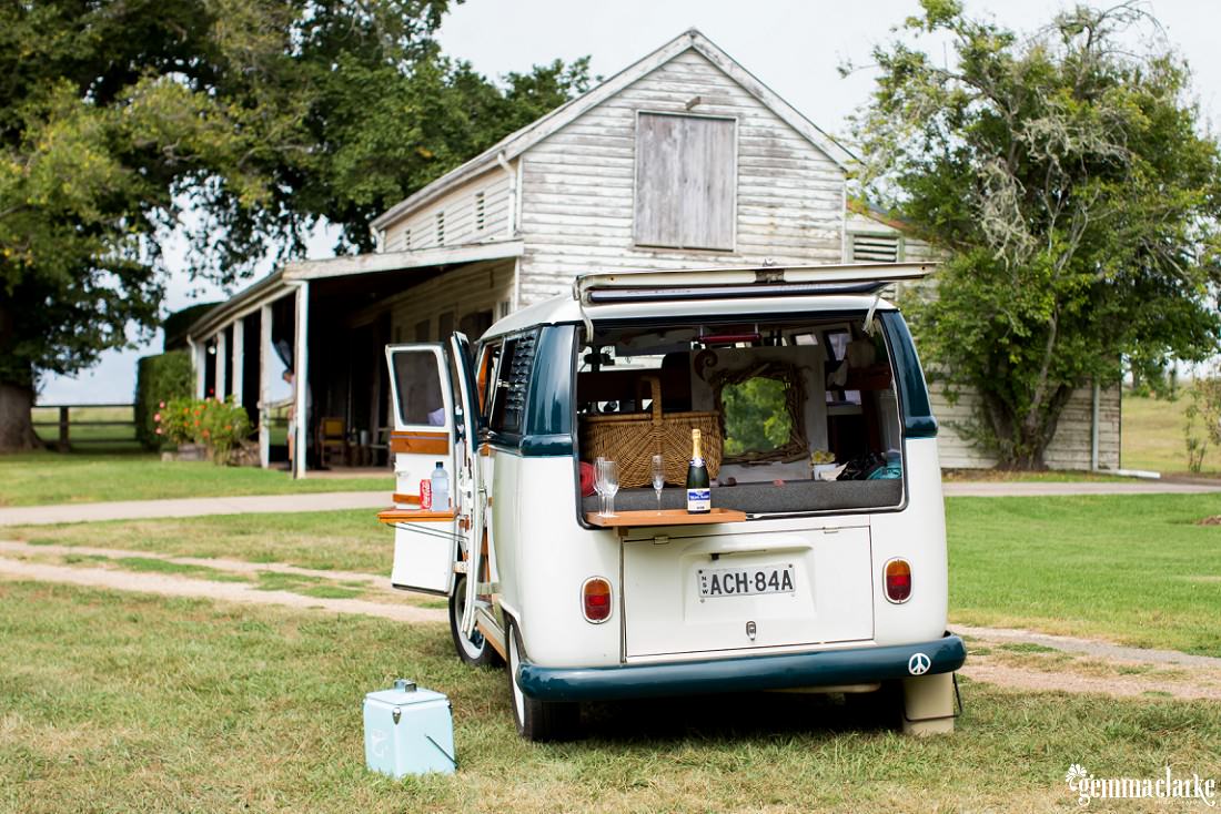 A Volkswagen Microbus with rear doors open and a small table with champagne and flutes, parked in front of a country house