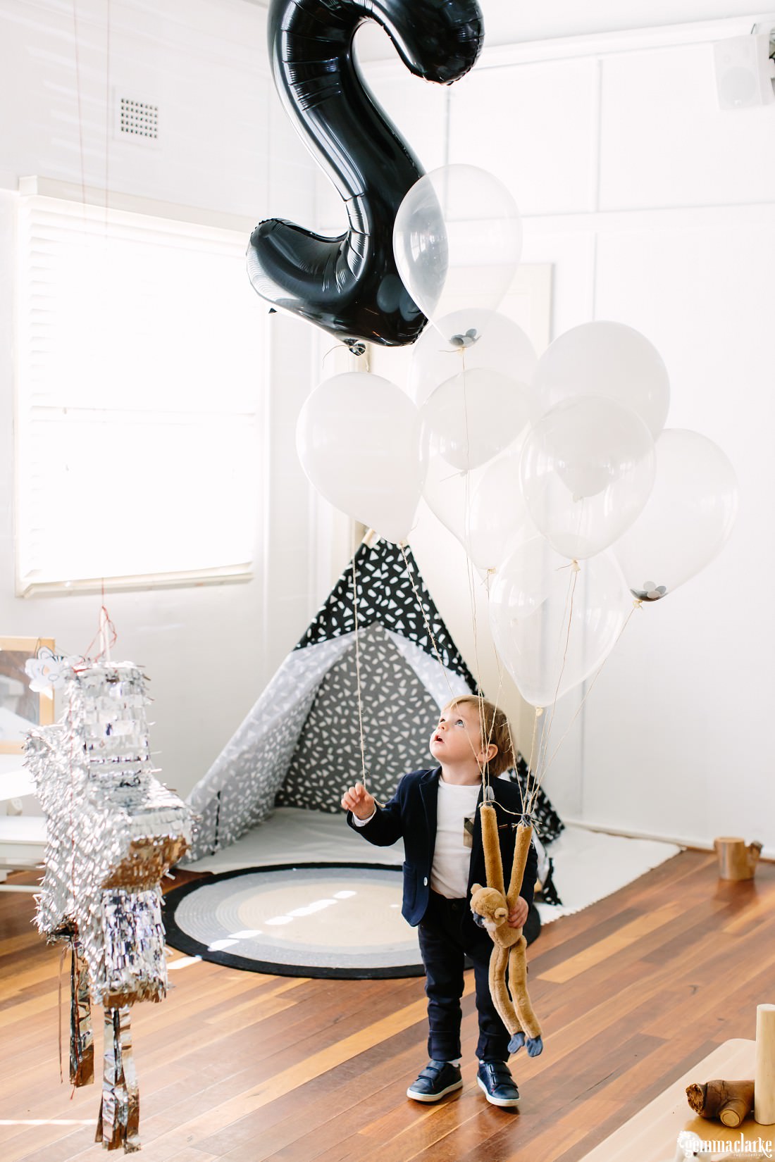 Henry stands in his room with a decorative tent behind him, the star shaped pinata in front of him whilst holding his large number 2 balloon and a light brown monkey toy which is attached to balloons and floating.