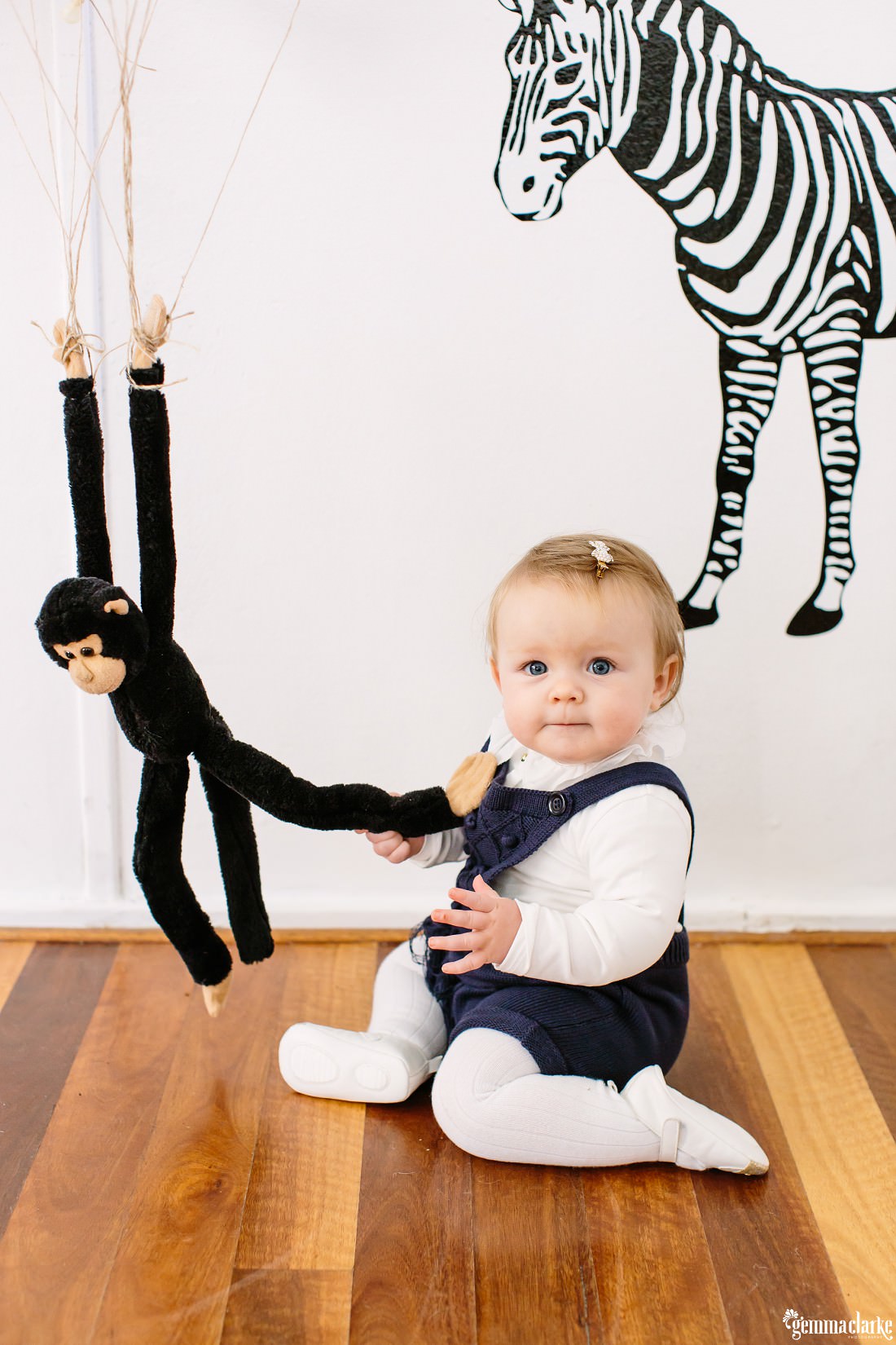 There is a toy monkey with long arms and legs that is attached to helium balloons which is making him float and Florence has grabbed his leg and smiles for the camera.