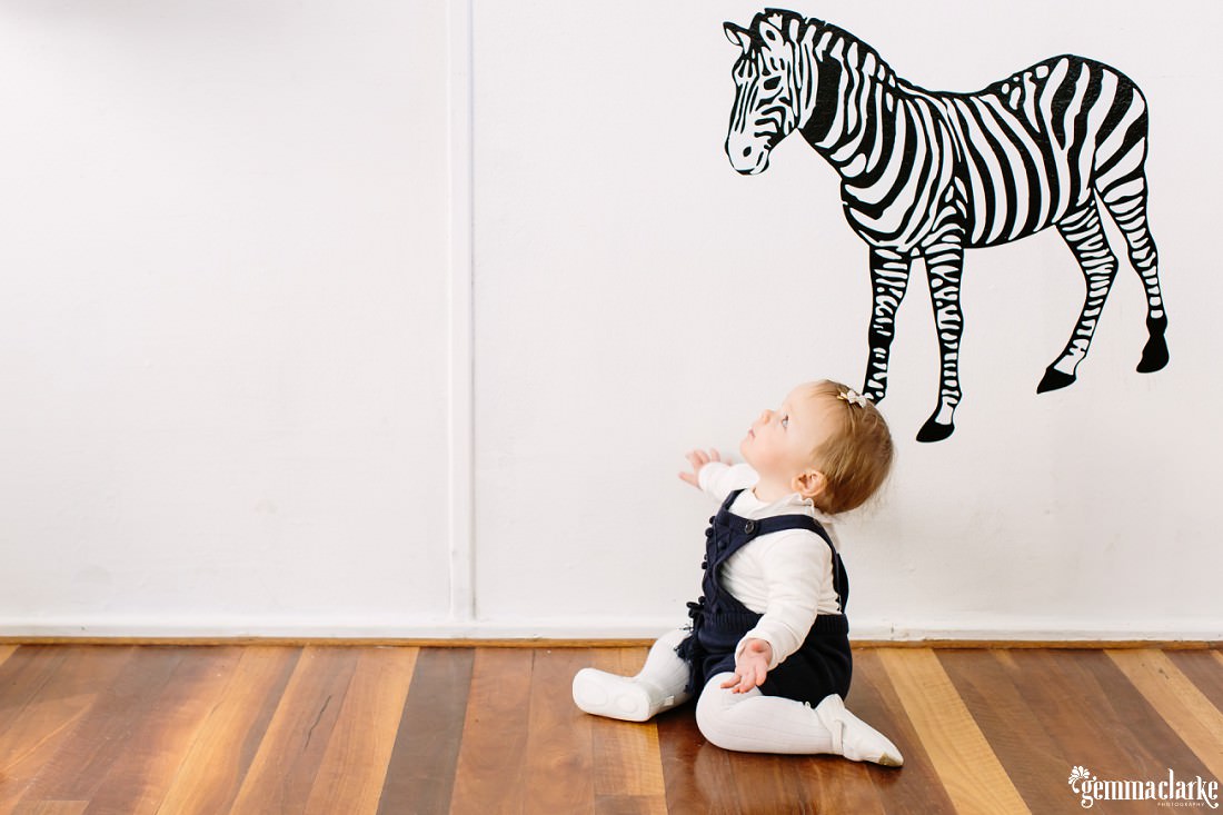 Florence is sitting on the wooden floor against a white wall with a zebra on the wall. She is looking up at the zebra.