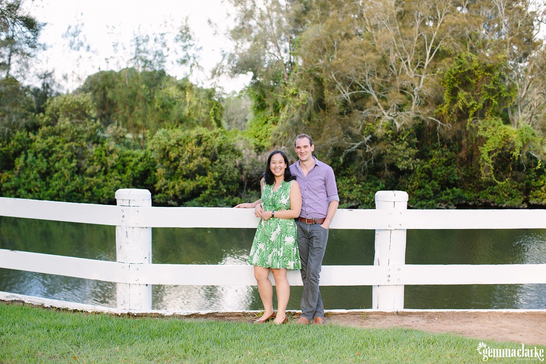 A couple standing in front of a white wooden fence with the lake in the background and overhanging trees - Couple Portraits in Bankstown